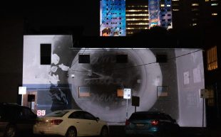 Gertrude Street Projection Festival. A negative black and white image of a clock is projected onto the side of a building. Cars are parked in front. Behind colourful projections appear on the side of tall blocks of flats.