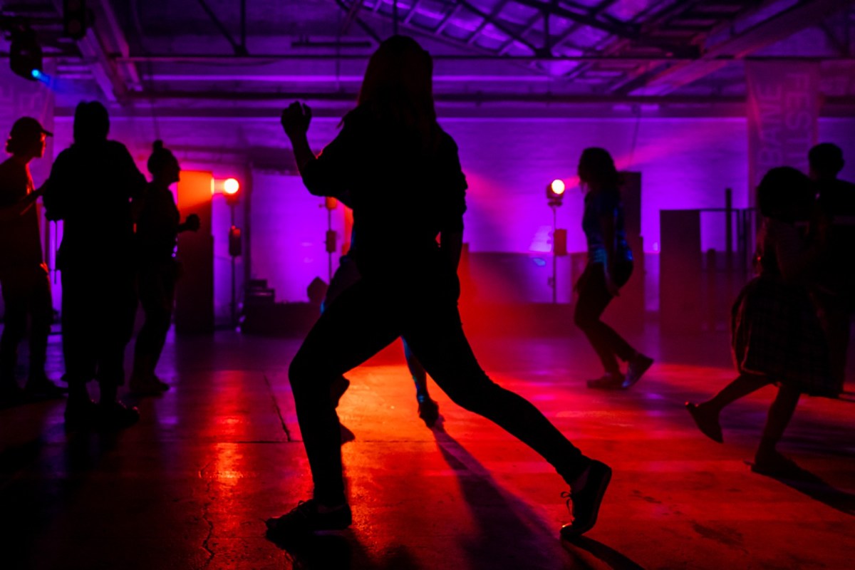 Dancers in a large warehouse space are silhouetted against purple and red lights.