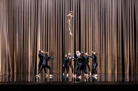 A group of people all dressed in black leotards are looking up, about to catch a single performer who is dressed in gold and is seen in mid-air. They are standing in front of gold drapery.