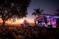 The 2023 Darwin Festival's closing weekend at the Ski Club. A crowd is gathered around a brightly lit stage at sunset. Palm trees and other trees are silhouetted against the sunset and festooned with strings of lights.