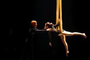 Two performers, one is a female opera singer with bright orange hair, and the other is a female acrobat hanging from silks, on a darkened stage. cross art form.