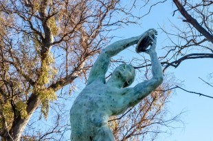 Bronze statue in park with blue sky and trees in background.