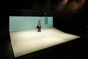 A person with dark trousers, short hair and a towel around their neck is standing in the distance of a set made to look like an empty pool. There is a stepladder on the top right. Swim.