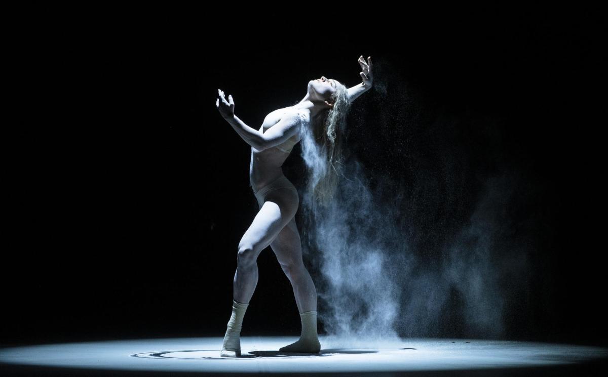 A single dancer is spotlit. She is all in white and covered in powder that falls off her as she poses.