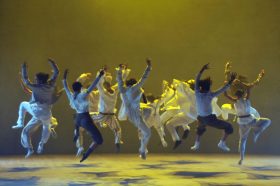 Dancing as fast as we can. A troupe of 12 contemporary dancers are all jumping in the air at the same time, with arms raised and bodies turned toward the back of the stage. They are mostly in white, with a couple in black trousers, and bathed in yellow light.