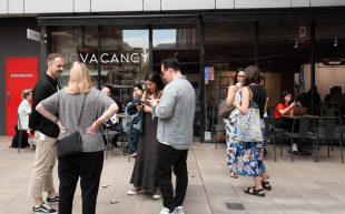 A crowd gathering outside No Vacancy. People standing outside with a glass window behind them with the words ‘NO VACANCY’.