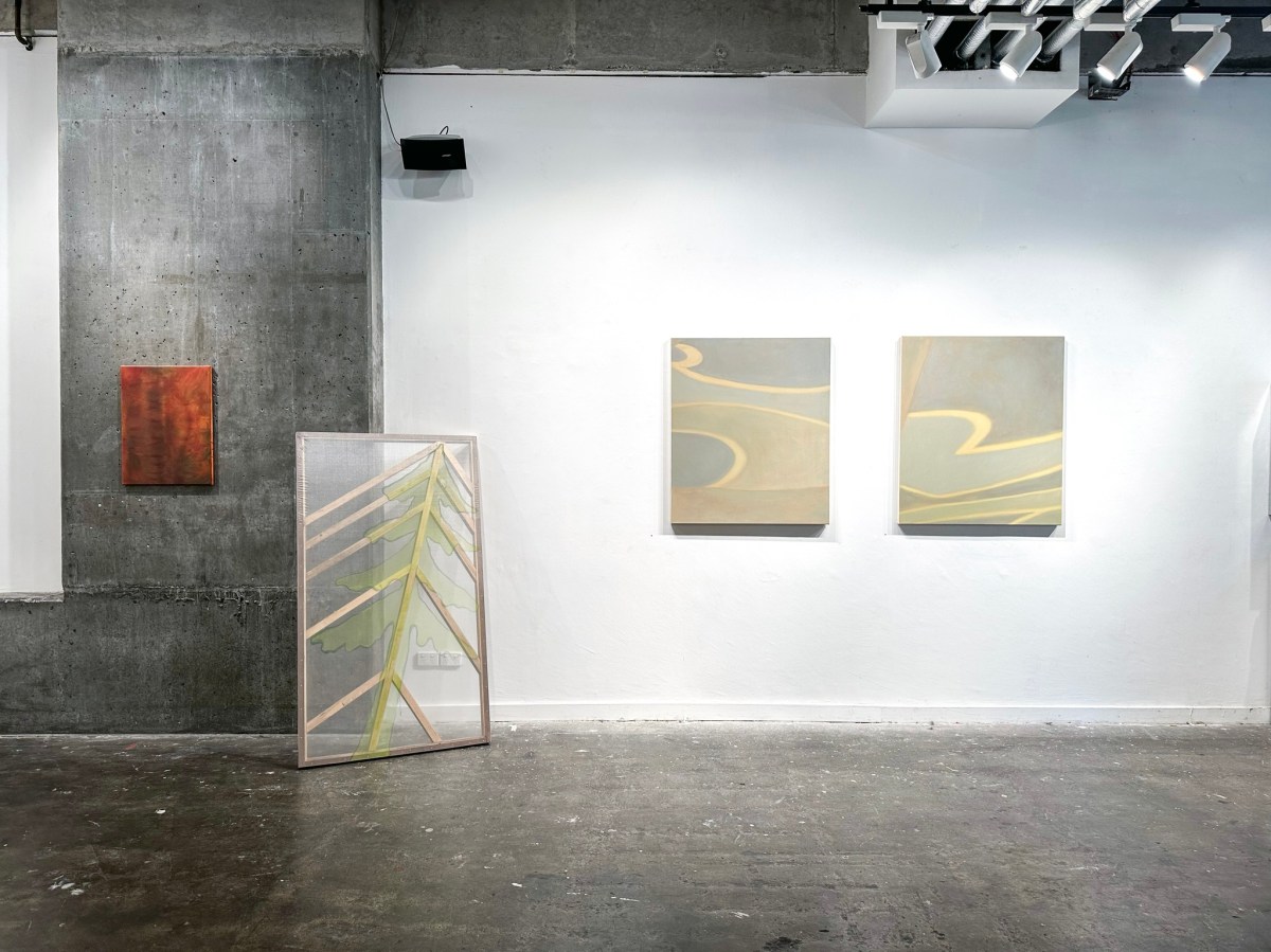 ‘The Softest Place On Earth’, installation view at No Vacancy 2024. A spacious gallery with exposed concrete floor and walls. One wall is painted white, with two paintings installed. The paintings have soft hues of blue, green and grey, and feature abstract swirls. To the left is an installation with a flat transparent screen on a timber frame with a tree-like pattern. On the far left is a smaller work installed on the wall, depicting red and orange hues.