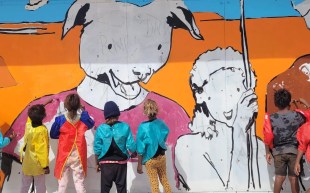 Youth participation in the Community Jam Wall (Mai Wiru Big Shop), lead by Warlpiri artist Robin Quinsten Jampijinpa Brown and visiting artist, Kaff-eine. A group of kids working on a colourful mural with their backs facing the camera. The mural depicts a dog and a human figure.