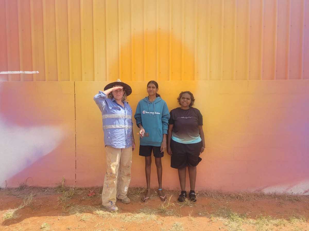 Artists Brontë Naylor (a woman with pale skin and blond hair wearing a purple shirt and cargo pants covered with paint), Selina Nakamarra Hunter (an young woman with brown skin and dark brown hair wearing a turquoise hoodie holding a paintbrush) and Mahalia Nampijinpa Hargraves (a young woman with brown skin and short dark brown hair wearing a tshirt and shorts) who did the Safe House community mural. They are standing in front of a wall painted in a gradient of yellow, orange and pink. 