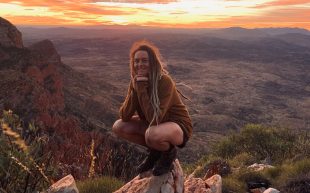 Imogen Batt-Doyle, winner of The Cloncurry Poetry Prize 2024 with an ode to the Outback. Photo: Supplied. A person squatting on the peak of a mountain with the sun setting in the background.