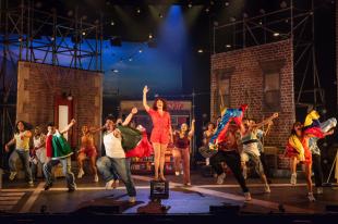 A woman in a short red jumpsuit is standing on a milk crate in the centre of a stage. She is surrounded by dancers all dressed in casual wear.