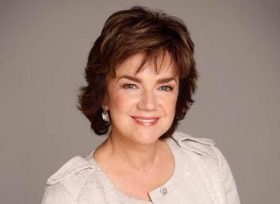 Actor Janet Andrewartha smiles for the camera in a publicity photo. She is an Anglo-Australian woman with carefully styled brown hair and wearing a white jacket.