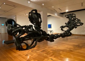 Black twisted wave like sculpture suspended from ceiling in gallery setting. Lucy Irvine.