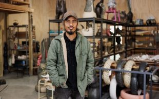 Mark Valenzuela, recipient of 2025 Porter Street Commission, in his studio. Photo: Rosina Possingham. A middle-aged man of Filipino descent with a shortly trimmed black beard smiling. He is wearing a brown cap with a green jacket and hands in his pockets. In the background is a studio setting with an array of objects on shelves.