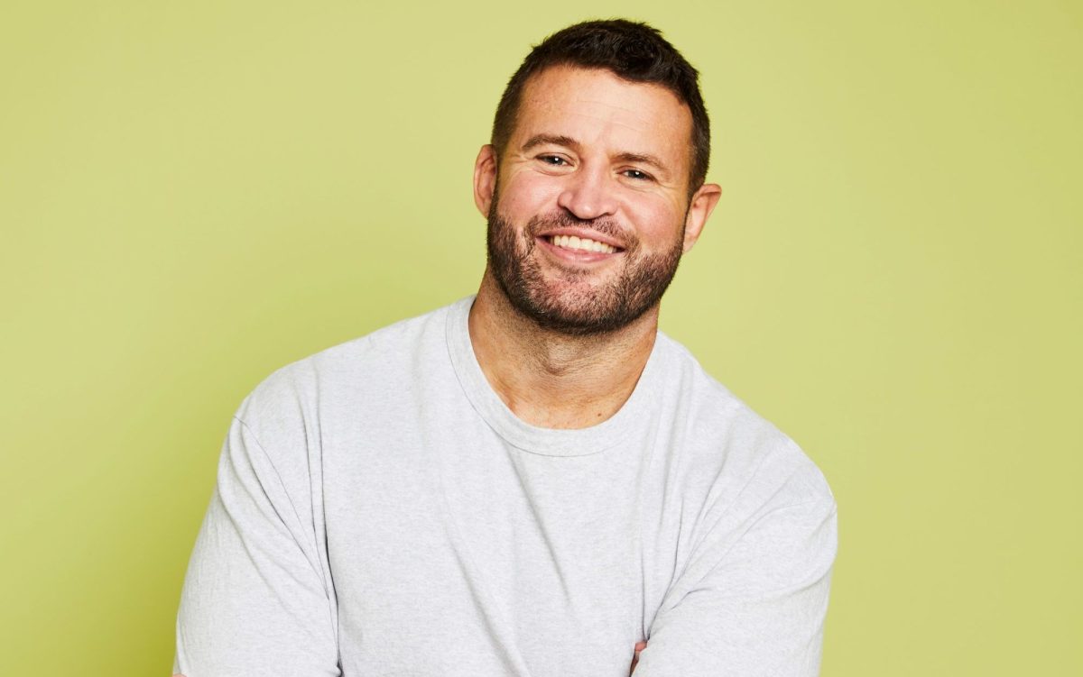 An Anglo-Australian man smiles affably at the camera. He has short dark hair, stubble, and wears a white t-shirt. He is standing in front of a light yellow backdrop. Andrew Hamilton.