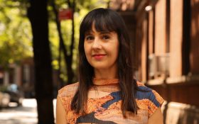 Melissa Loughnan, the newly appointed Fair Director at Melbourne Art Fair for 2025 and beyond. Photo of a middle-aged woman with pale skin and long brown hair with straight bangs. She is smiling at the camera, wearing an abstracted orange and navy sleeveless top on an outdoor residential footpath.