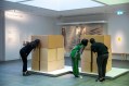 Three people looking inside cardboard box structure in Museum. Models.