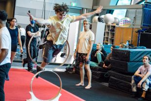 A young circus artist spreads their arms and smiles as they dive through a vertical ring. Other young people watch on in the circus training session.
