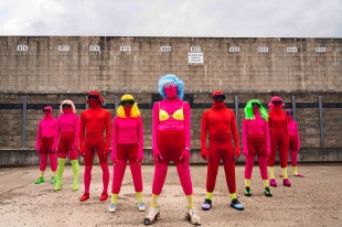 A group of red leotard clad performers stand in formation in front of a wall with their faces also covered, but colourful adornments like wigs or bras breaking up the red.