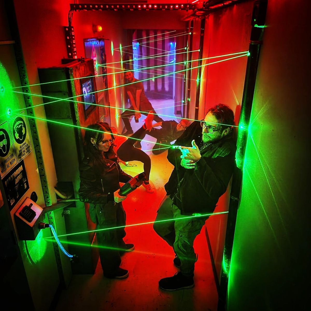 Several young people stand or lie in a corridor leaning against walls and surrounded by red light and green laser beams.
