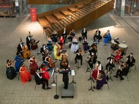 A string orchestra playing in the atrium of a large retail space in front of a staircase. The musicians are masked and wearing black tie dress, with many colourful dresses included.