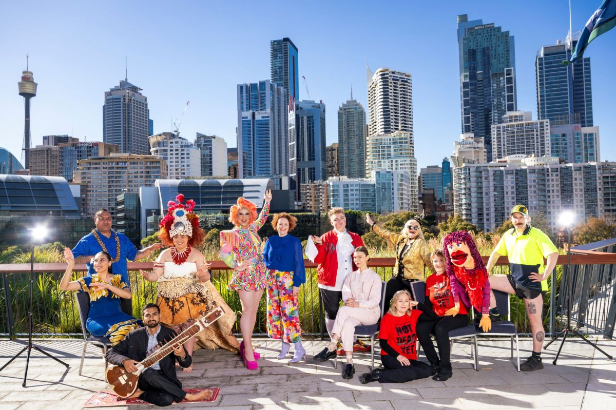 A colourful group of Sydney Fringe performers, including a drag queen, a puppet and puppeteer and others, pose with the Sydney CBD skyline in the background.