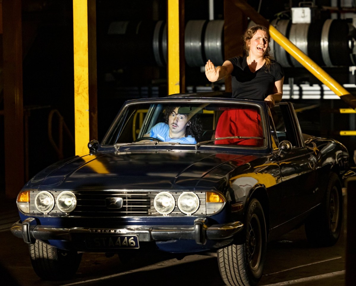 A theatrical scene of two young people in a dark blue Triumph car. A man in a blue T shirt is driving, while a woman in red pants and black shirt is standing with her head out of the sunroof screaming.