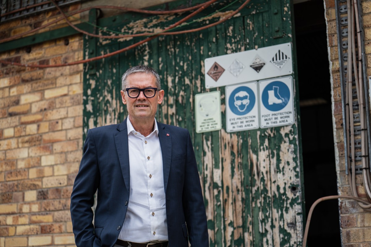 A middle aged Caucasian man with grey hair and black framed glasses, white shirt and suit jacket  stands in front of a paint peeling doors with industrial signs on it.