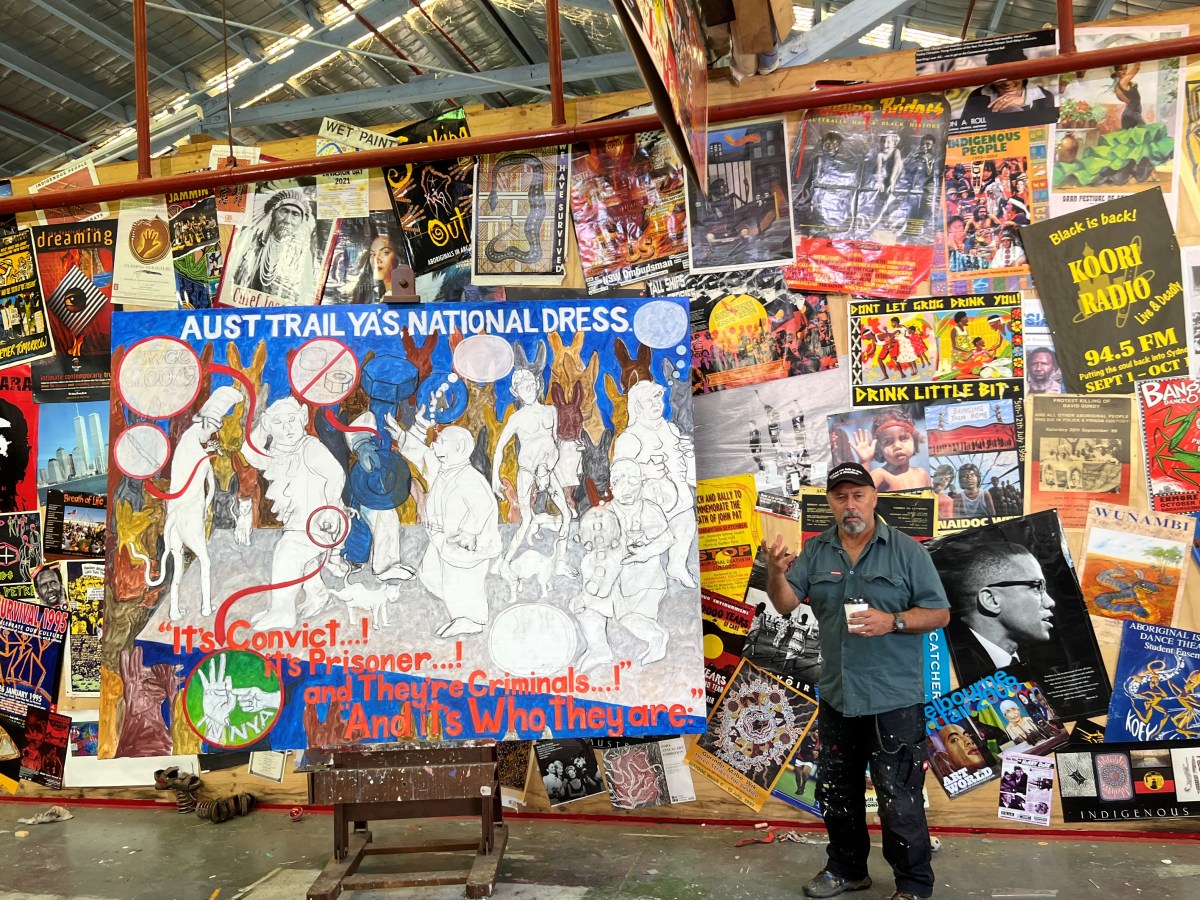 An Indigenous artist stands in a studio in front of a large work and a wall covered with political and socially aware art works.