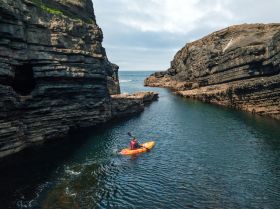 A man in a red life jacket paddles a bright orange kayak across a rippled stretch of water between two rocky cliffs.