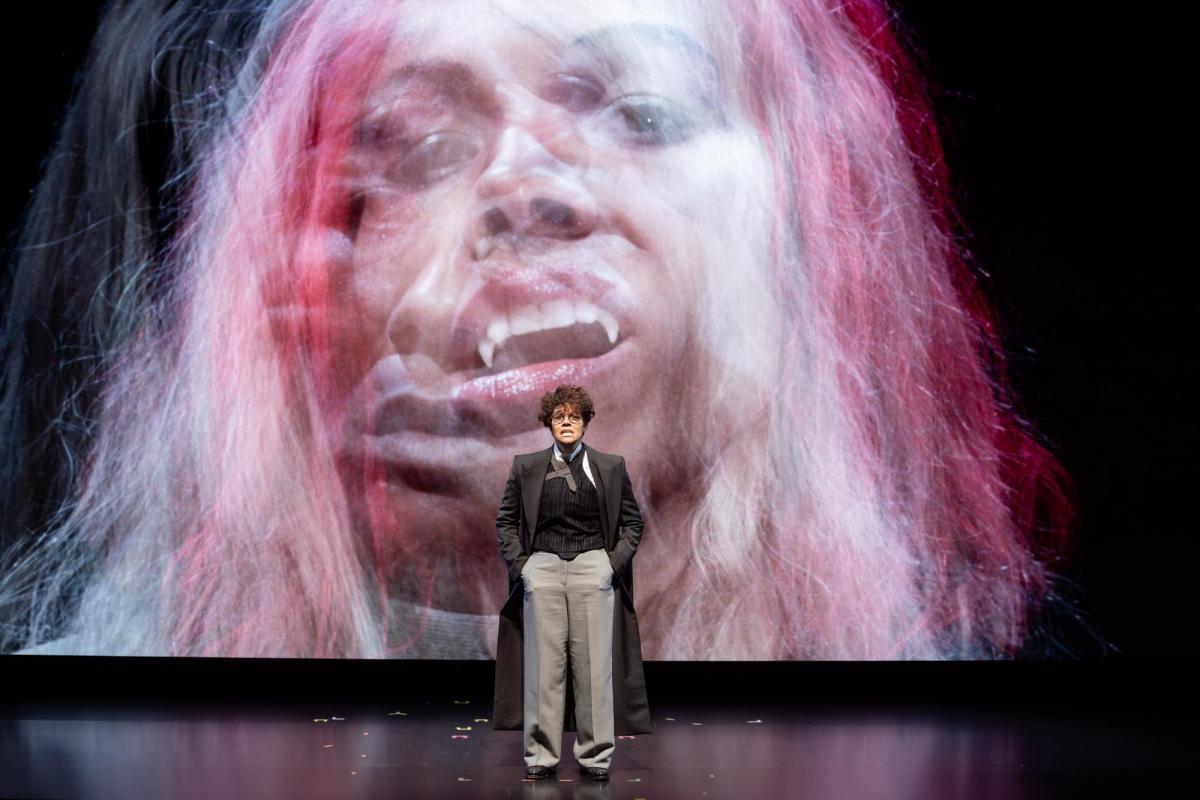 A woman dressed in pants and a long coat is standing in front of a large screen with an image of a close up headshot of a fuzzy pink and long-haired creature with fangs.