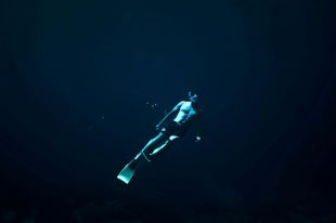 A moody photograph of a male diver underwater. His shirtless torso, facemask, snorkel and flippers are brightly lit against the deep blue ocean background.