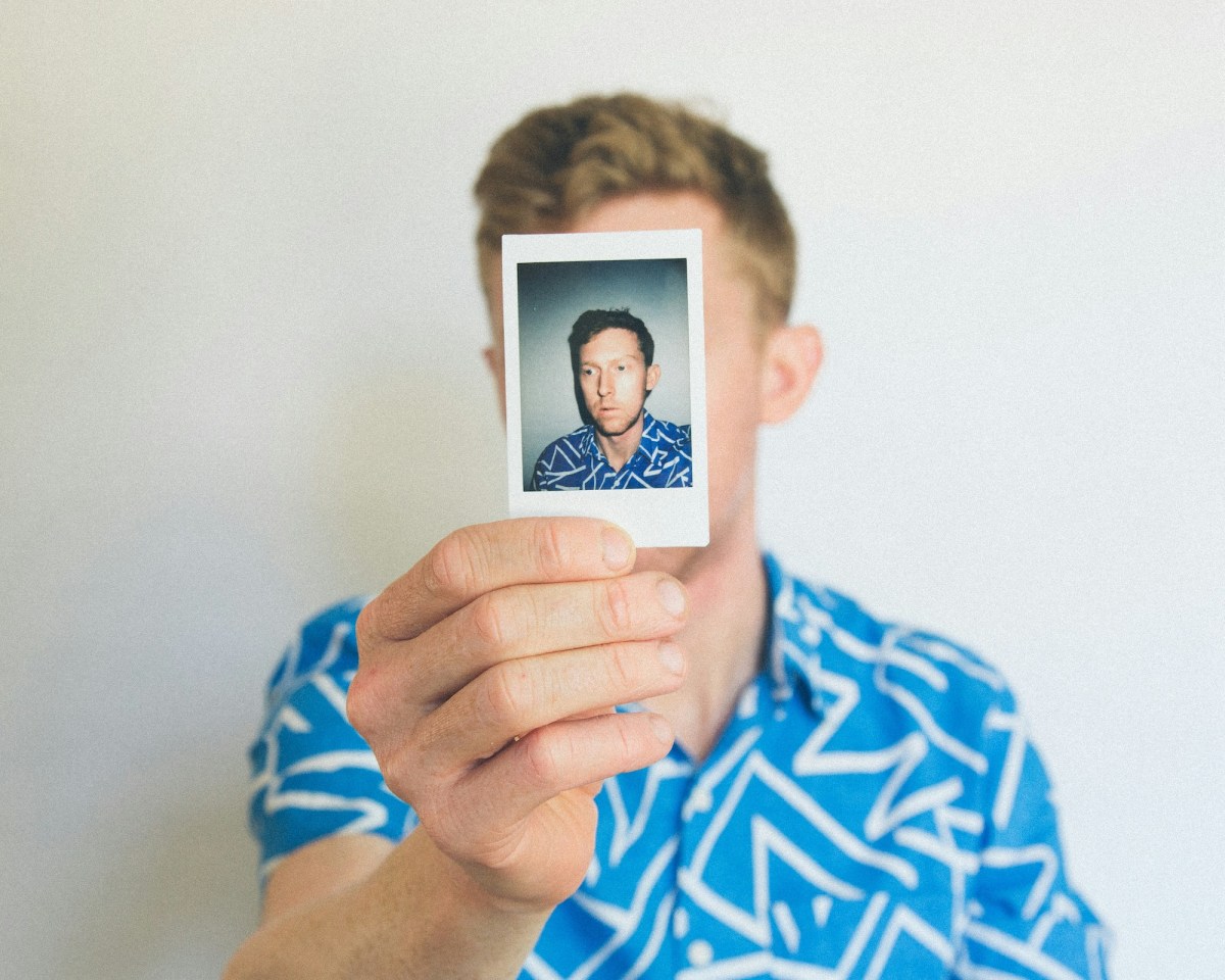 A Caucasian man in a short sleeved blue shirt with geometric white lines on it holds up a snap of himself obscuring his face.