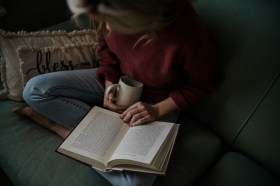 Woman in jeans and red jumper reading a book inside with warm drink.