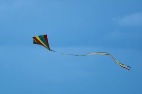 A colourfully-striped triangular kite with a long, bright tail flies against a clear blue sky.