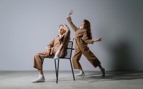 Pursuing a career as a dancer comes with financial burdens and an uncertain future. Two dancers wearing light brown workwear onesies in a white space. One is sitting with one foot propped up on the chair, their hand on their head, and another is standing behind in an elegant stance, extending their arms and one leg.