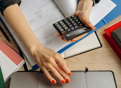 A white woman's arm and hand reaching over papers to work on a laptop, while she holds a calculator in her other hand.