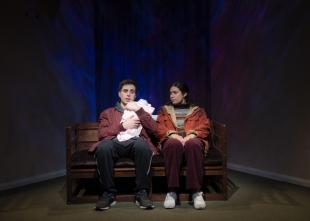 A young couple are sitting on a park bench The male is cradling a white bundle, made to look like a baby is within it.