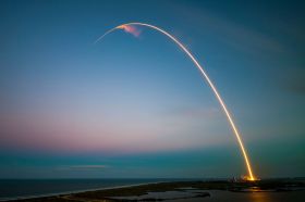 A time lapse photograph of a rocket's trail traced in a fiery arc from the launchpad against the darkening sky.