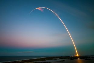 A time lapse photograph of a rocket's trail traced in a fiery arc from the launchpad against the darkening sky.