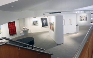 Stanthorpe. A white walled gallery space with art on the walls, pictured from the mezzanine level looking down some stairs.