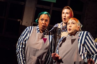 Two Remain. Three women are singing. They are wearing blue and white concentration camp stripes underneath beige aprons and head scarves.