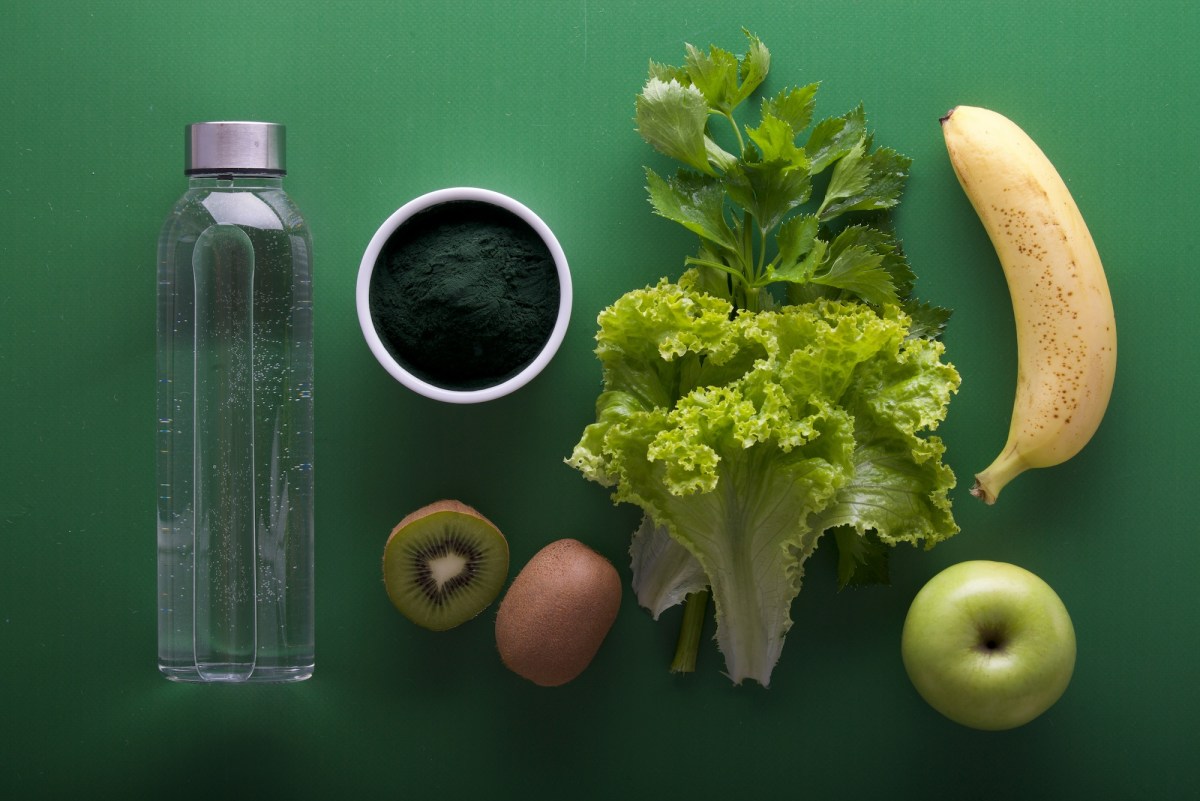 A green background with healthy food laid out - banana, kiwi, apple leafy greens and bottle of water.