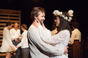 A man and a woman are wearing bridal white. She has a crown of flowers on her head. There are other people in the background talking.