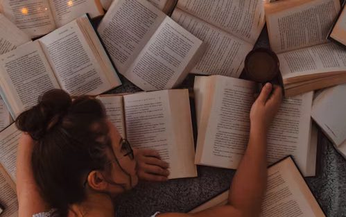 A brunette woman is lying face down on a pile of books with their pages splayed open.