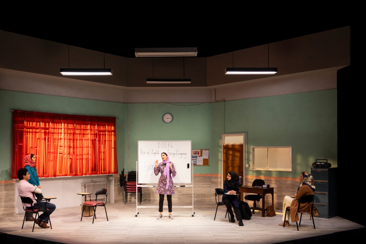 English. Southbank Theatre. A classroom set up on stage, with pastel green walls and an orange curtain on the left side. A woman is standing in front of a whiteboard. There is a man and three women surrounding her.