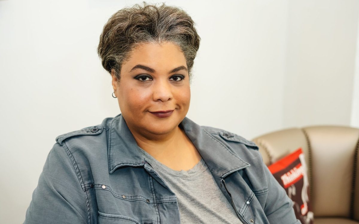 Roxane Gay is a woman with light brown skin, short pepper hair. She is looking at the camera with a confident smile and an arch in her brow, sitting on a tan leather sofa against a white wall.