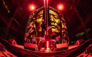 Gabber Modus Operandi performing at Royal Exhibition Building as part of Now or Never 2024. A performer is at the front of a stage wearing puffy sleeves and a face cover, with a large-scale projection of a human face behind. The stage is aglow in red light.