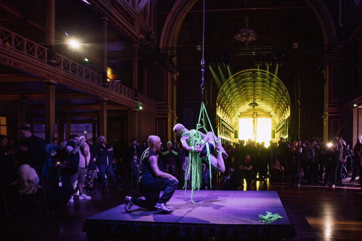 Live performance by Luke George at Royal Exhibition Building on 22 August. A small crowd gathers around a slightly elevated platform where two human figures are visible. One is kneeling while the other is tied up in neon green bondage and hovering above the ground. On the far side of the photo is the arch of the Great Hall leading into the main performance area, cast in a bright yellow glow. 