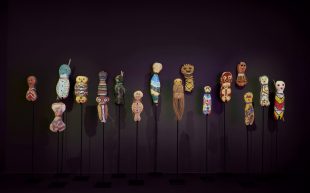 A collection of 17 small figures with circular heads and bodies, painted in bright pigment and displayed in front of a black wall at AGNSW.